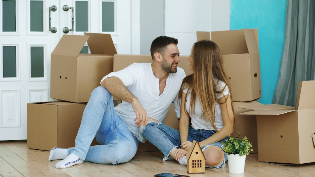 A smiling couple takes a break from unpacking, sitting together on the floor of their new, bright living room surrounded by boxes.